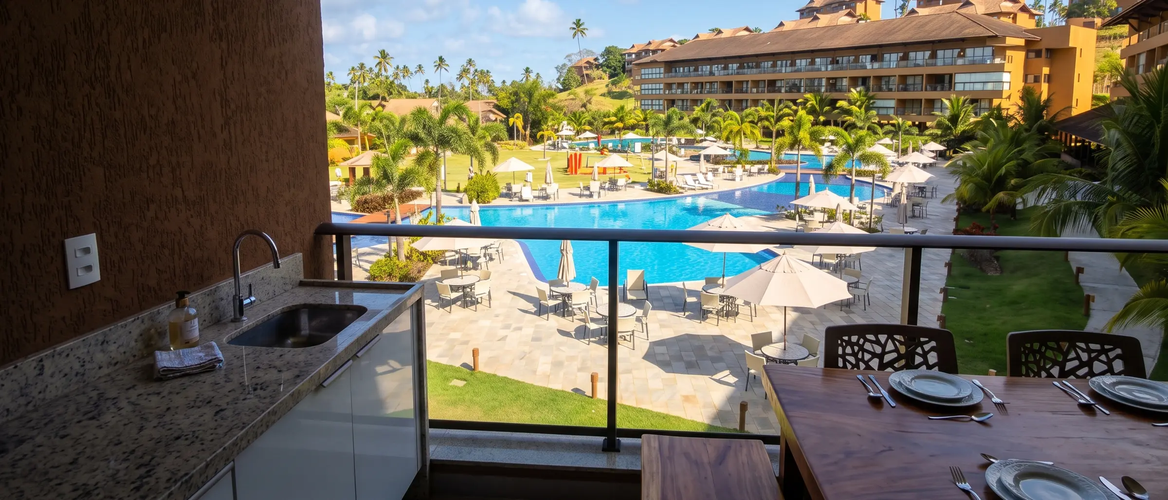 Varanda do flat com mesa posta, pia de apoio e vista para as piscinas do condomínio